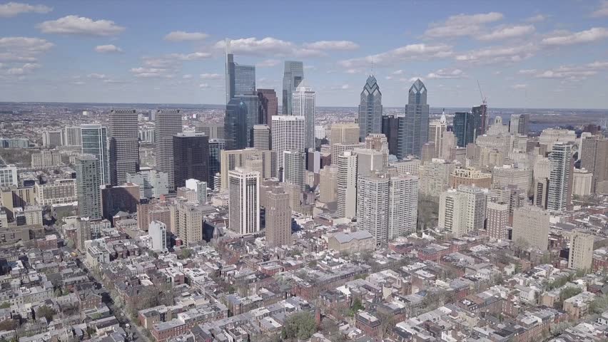 Dynamic drone shot of Philadelphia downtown architecture featuring Liberty Place and Comcast Center. Cinematic panoramic footage over urban landscape with dense housing and blue sky horizon.