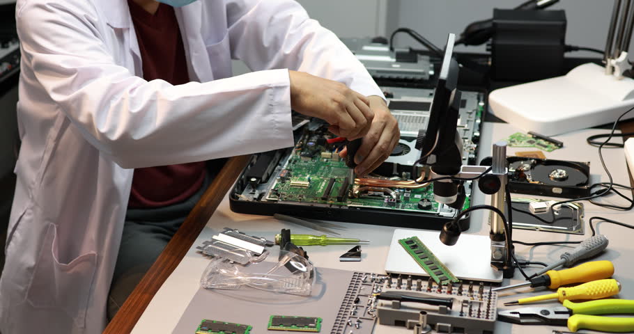 Technician in a lab coat repairing a computer motherboard at a cluttered electronics workbench with tools, memory modules and hard drives.