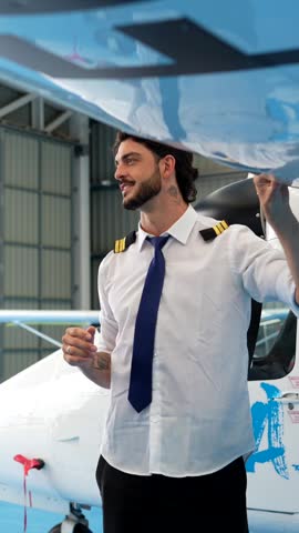 Handsome male aviator with a beard wearing a pilot uniform standing under the wing of a small airplane inside a hangar, confidently inspecting the aircraft and talking about its features