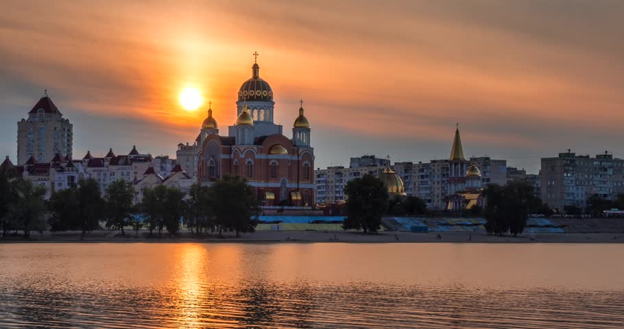Golden sunset over Obolon Embankment in Kyiv, Ukraine. Warm evening light illuminates a riverside church and city skyline reflected in the calm waters of the Dnipro River.