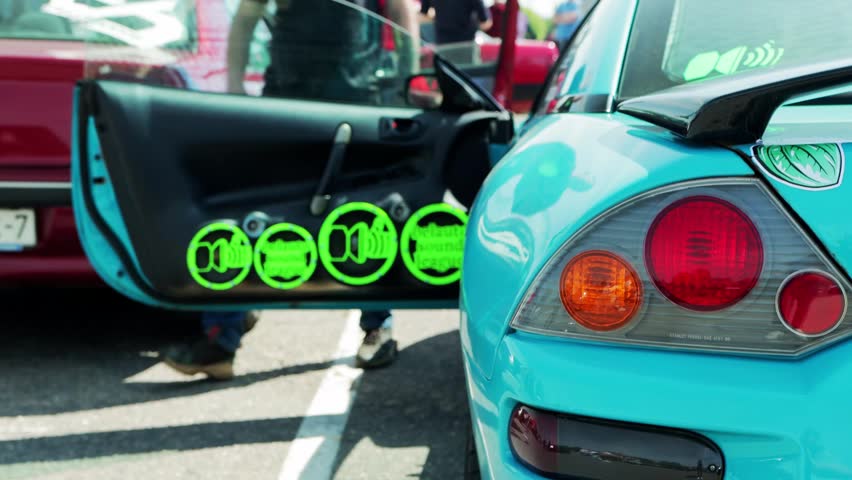 Close-up view of back of flashy custom blue sport car displaying bright red and orange tail lights. Open door reveals modified interior with vibrant green circular sound system speaker grilles