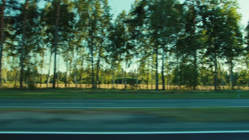 Dynamic view from side window of moving vehicle, capturing blur of green trees and sunlit summer sky. Freight trucks and passenger car pass by on multi-lane road.