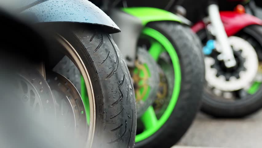 Focus on front section of blue, green, and red motorcycles side by side. Water droplets visible on fender of blue bike. Disc brakes and tire treads are prominent. Blurred background suggests outdoor s