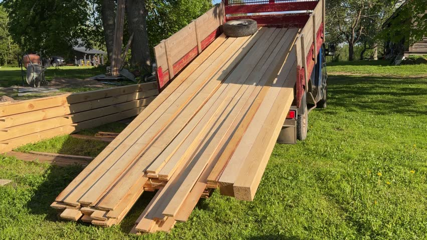 A truck is unloading timber cargo in a countryside yard. The footage shows how truck is lifting the cargo using hydraulics and timber is falling on the ground. 