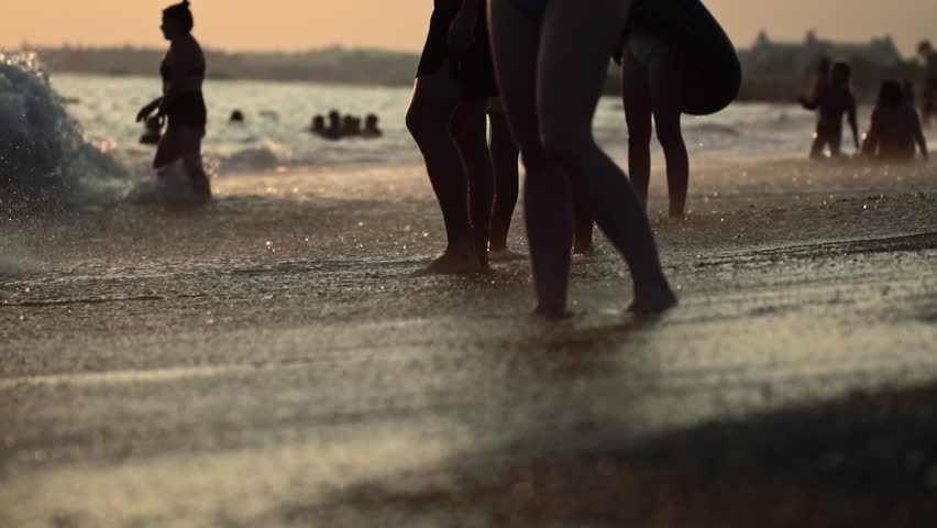 Lower body shot of people standing in shallow sea water interacting with gentle waves on beach at sunset. Golden light reflects on water surface, creating shimmering effect. Vacationers enjoy peaceful
