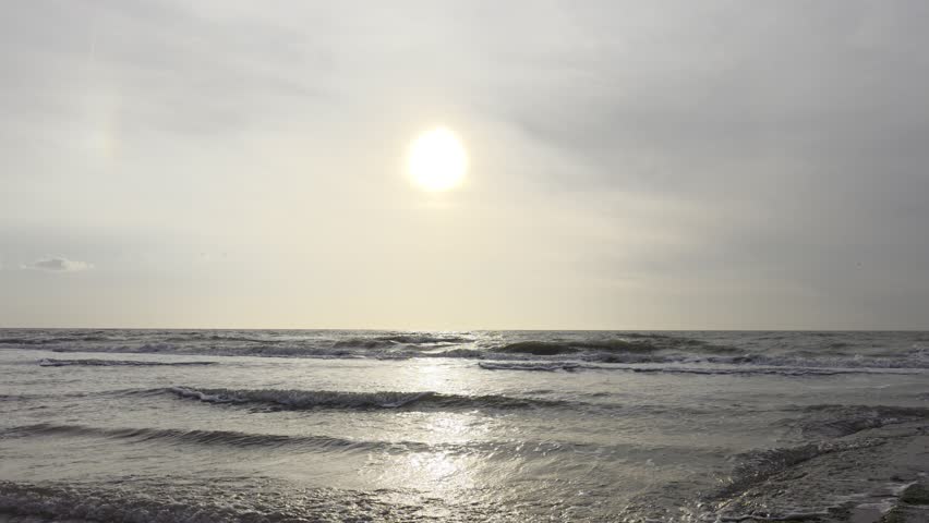 A scenic view of the bright sun shining through cloudy sky over calm ocean waves moving towards the shore