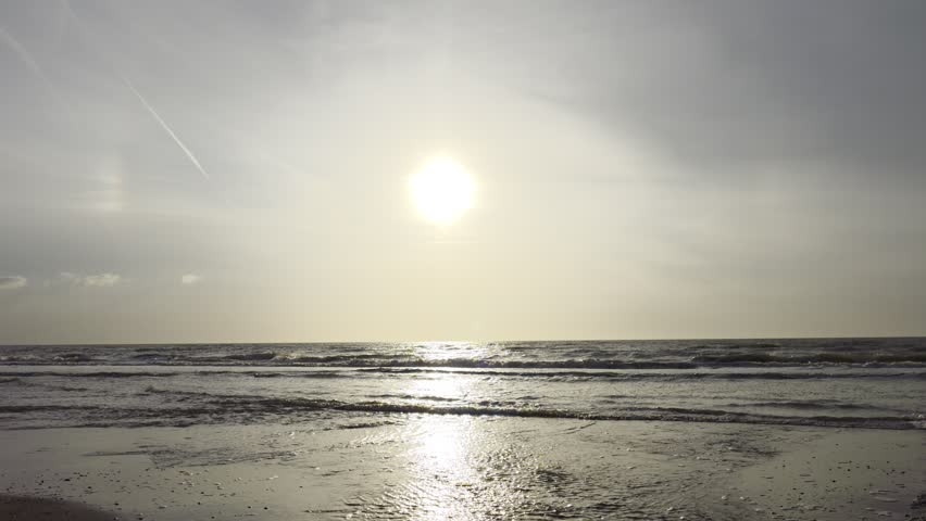 A scenic view of the bright sun shining through cloudy sky over calm ocean waves moving towards the shore