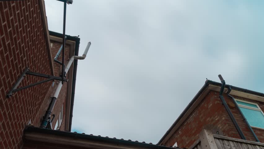 Dark cloud timelapse drifting between residential brick houses. Slider shot narrow urban courtyard under overcast sky. Moody neighborhood architecture framing fast moving clouds atmospheric weather