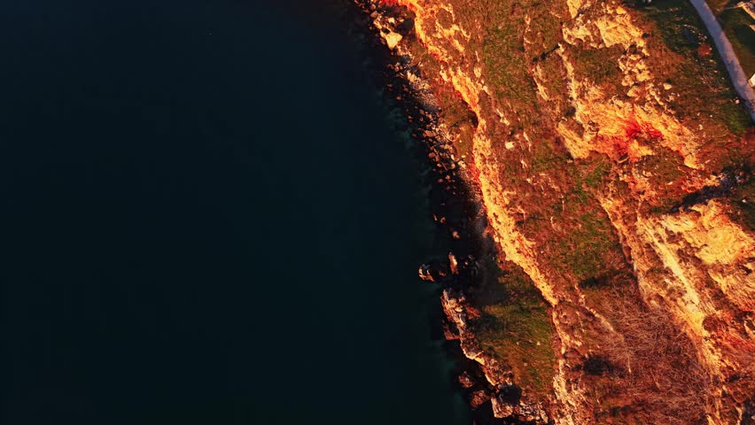 The sun sets at the edge of the coastline, casting shadows on the rocky shore. The calm water meets the land, creating a boundary between elements at dusk.