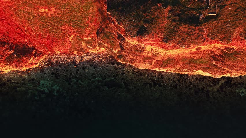 Aerial view shows a rocky shore at sunrise with bright colors glowing along the land. Clear water meets the dry land near the rocky edge. Nature appears vibrant and inviting.