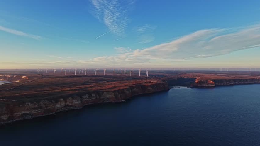 Wind turbines stand on a cliff near the water. The sun sets on the horizon, casting warm light on the landscape. A clear sky shows scattered clouds above the area.
