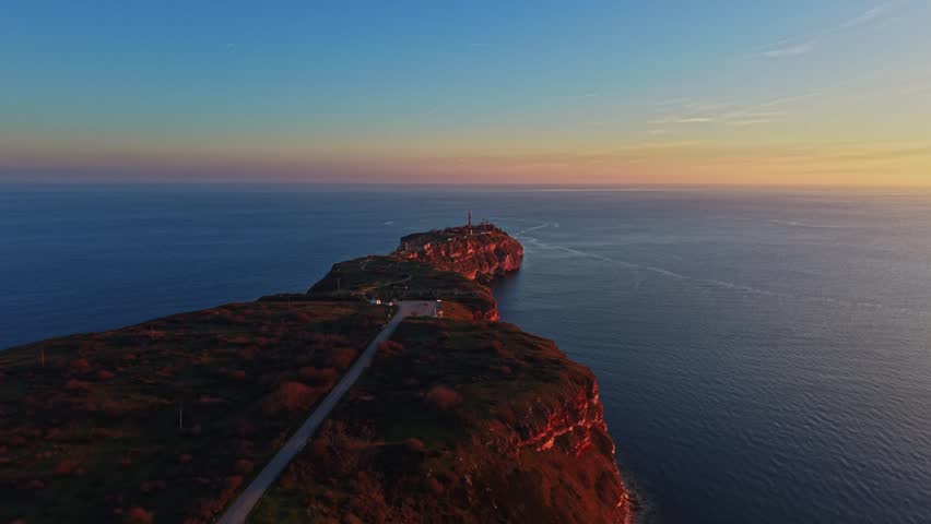 The scene shows a rocky island surrounded by water at sunset. The sky is clear and the sun casts colors on the island. Waves gently move around the shore.