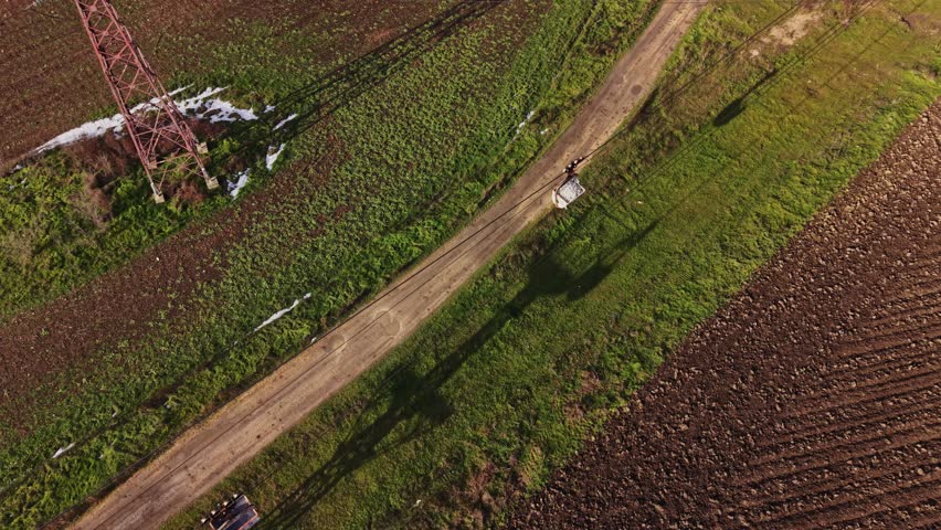 Two vehicles are parked on a dirt road in a field. A power line tower stands nearby as the sun sets, casting long shadows across the green grass and brown soil.