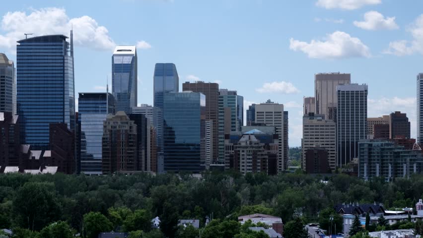 Scenic aerial view of Calgary city center featuring modern architectural skyscrapers. Dense urban skyline stands against blue sky with white clouds. Lush green trees fill the foreground park.