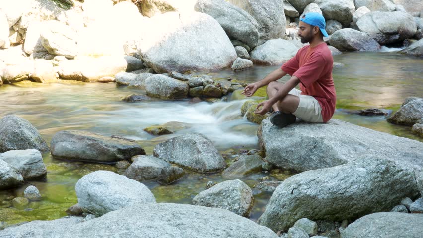 A man meditates peacefully on a rock beside a fast-flowing river in a scenic mountain landscape.