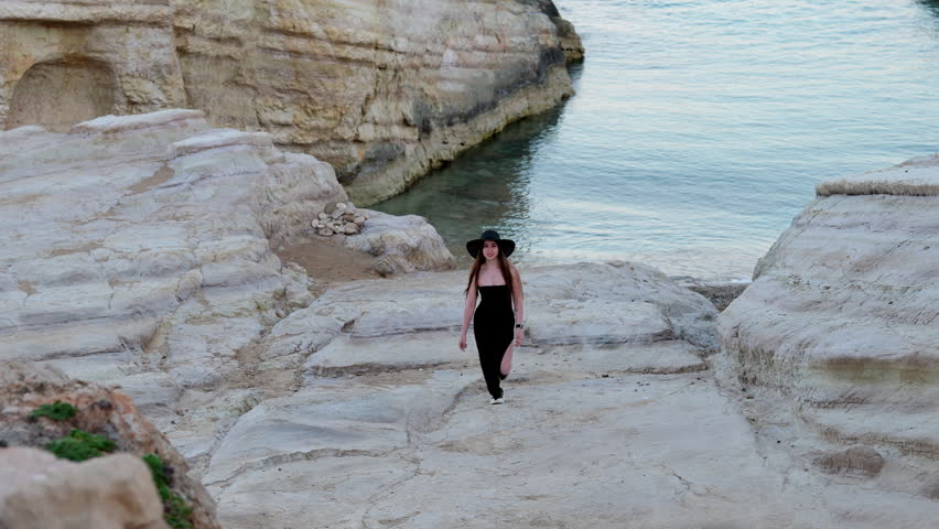 Young gorgeous woman in a long black dress and hat gracefully walking towards the camera along a beautiful rocky beach on a calm evening