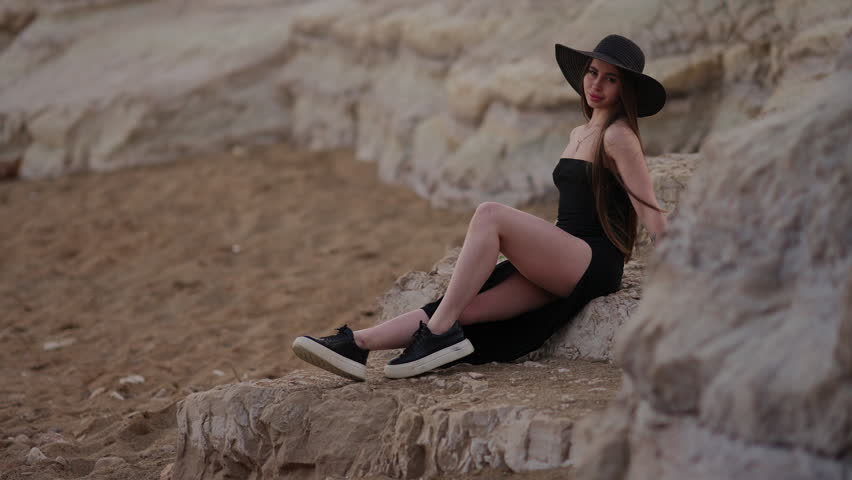 Stylish young woman with long hair wearing a black dress and hat posing for the camera while sitting on rocks at the beach