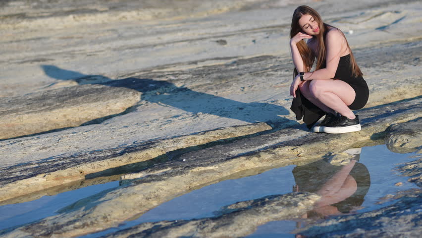 Beautiful young woman with long hair posing in a black dress while crouching on a rocky shore next to a puddle, reflecting her image