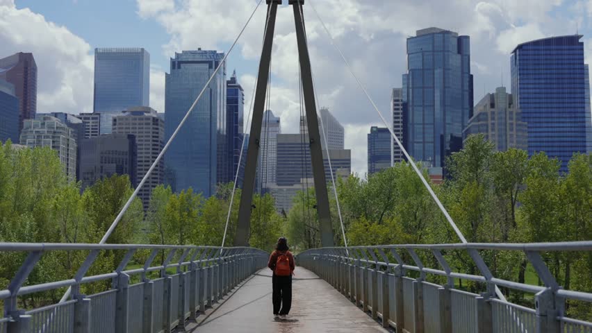 Lone figure crosses footbridge, modern skyscrapers in background, greenery, urban park pathway, daytime scene.