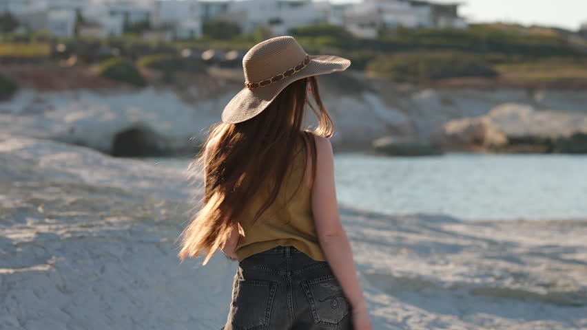 Cheerful young woman with long hair and a straw hat spinning and walking on white cliffs by the sea, enjoying summer vacation at sunset