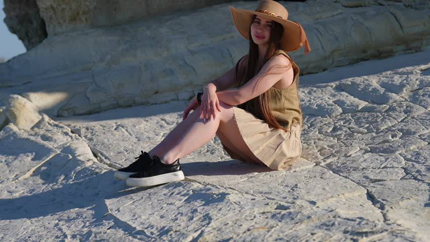 Beautiful young woman with long hair wearing a sun hat and a dress sitting on white volcanic rocks, enjoying a sunny day outdoors