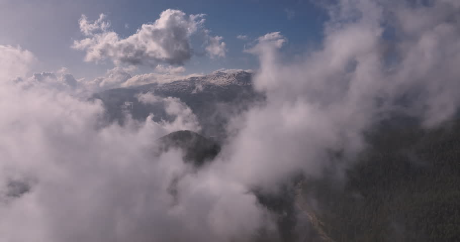 Aerial flight through misty clouds revealing snow capped mountain peaks and forested valley below in soft sunlight