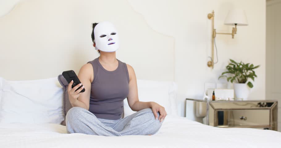 Woman wearing white mask is raising smartphone from lap to eye level on bed, preparing selfie. Facial, skincare, selfcare, wellness, bedroom, minimalist, neutral