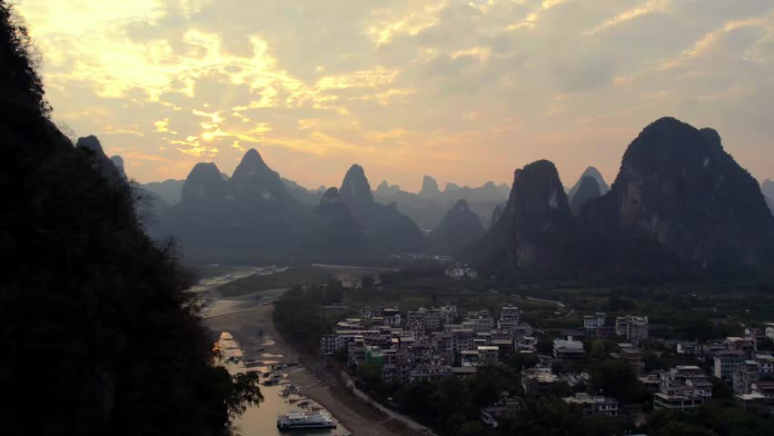 A dusk landscape featuring karst peaks, a riverside settlement, and a winding river under a softly lit sky.