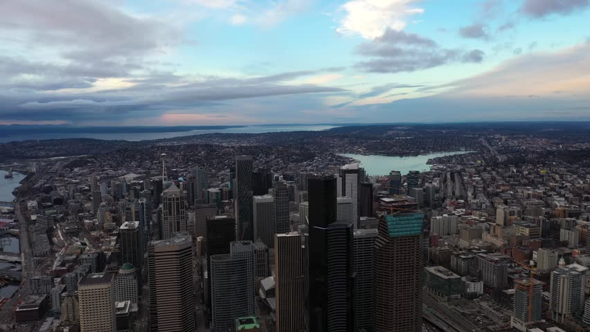Dynamic aerial footage gliding over downtown Seattle city center. Panoramic view includes towering buildings, Columbia Center, Lake Union, and Elliott Bay under a soft blue sunset sky backdrop.