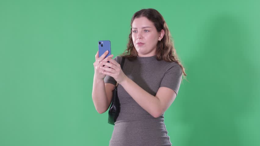 Young woman stands against green screen, holding smartphone up to take photo or scan QR code