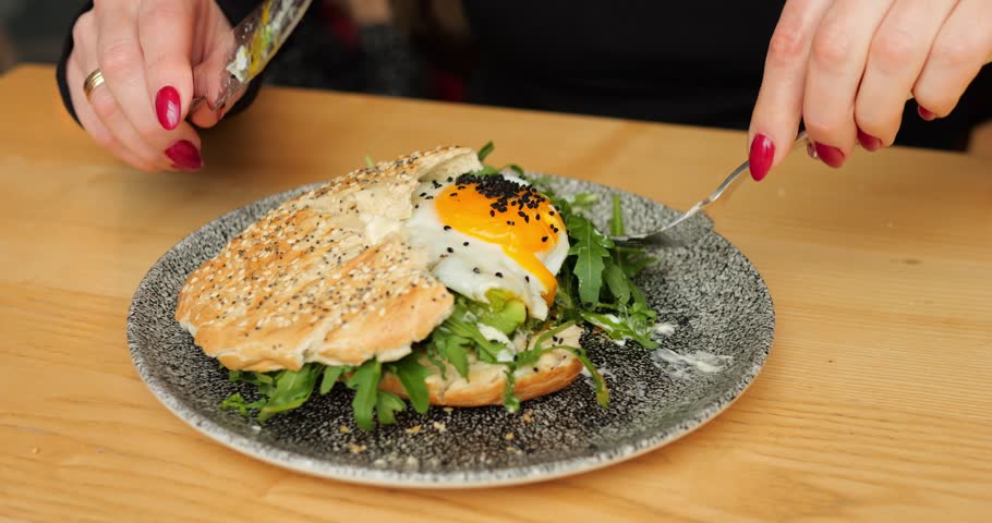 Diner spears arugula with fork and lifts fresh leafy forkful toward mouth. Knife steadies bun and yolk glistens on plate while person prepares next bite