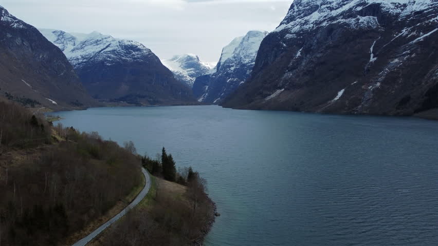 Curved road along a lake in Norway with snow-capped mountains and cloudy sky.