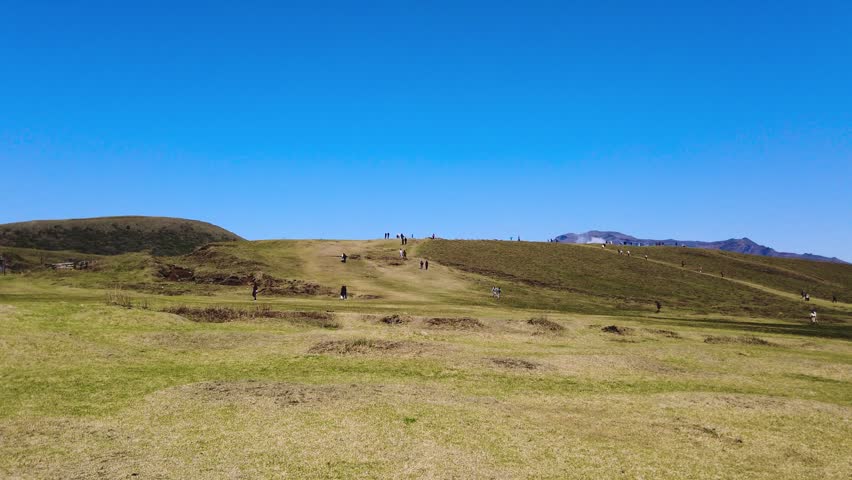 Wide shot of Kusasenri plateau at Mount Aso in Japan during spring season. People walk along dirt paths on fresh green hills under a clear blue sky in April. White smoke rises from active volcano crater in background. Scenic panoramic view of a famous volcanic tourist destination in Kumamoto.