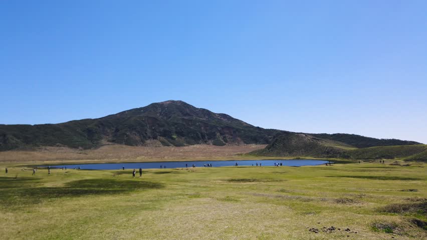 Kusasenrigahama volcanic plain inside Mount Aso caldera in Kumamoto Japan. Wide panoramic view of grasslands and fresh green fields with a pond during spring in April. Group of tourists and visitors walking around the crater area under a clear blue sky. Rural scenic nature landscape of volcanic tourist attraction on Kyushu island.