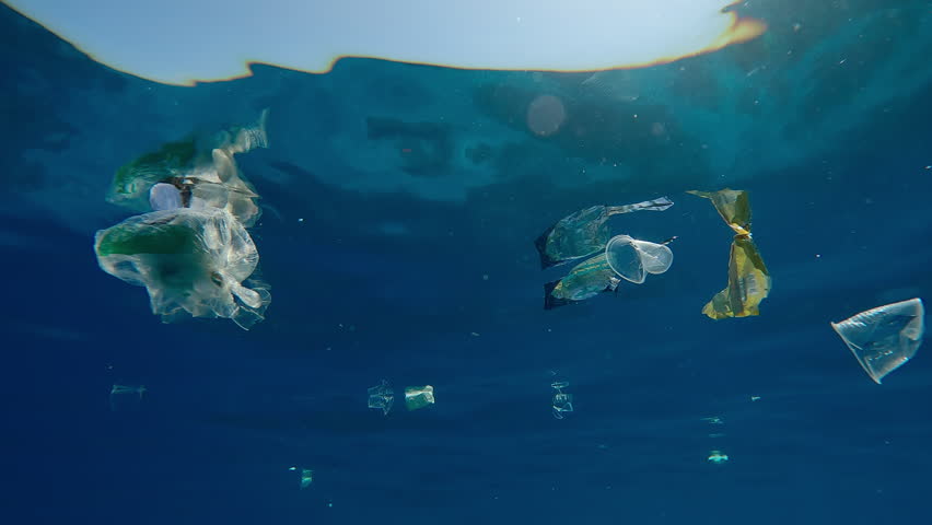 View from below on lots of of mixed plastic waste swims under the waves in dark blue clear water mirrored on its surface, in the bright sun glare, plastic pollution of the ocean, underwater shot