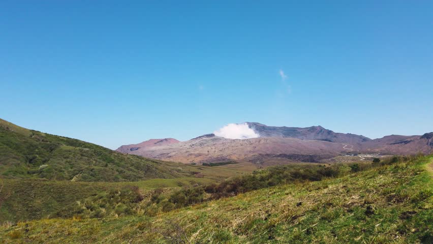 Panoramic view of Mount Aso volcano with smoke emitting from the active crater in Japan. Green grassy hills of Kusasenri plateau stretch across the landscape under a clear blue sky. Natural scenery showcases the unique volcanic environment during the spring season in April. Peaceful wilderness landscape captured on a sunny day highlights rural tourism destinations.