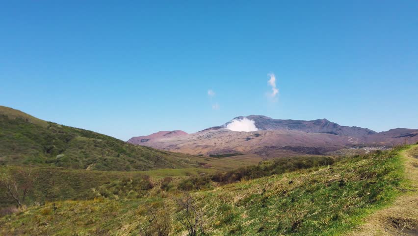 Panoramic view of Mount Aso volcano with smoke emitting from the active crater in Japan. Green grassy hills of Kusasenri plateau stretch across the landscape under a clear blue sky. Natural scenery showcases the unique volcanic environment during the spring season in April. Peaceful wilderness landscape captured on a sunny day highlights rural tourism destinations.