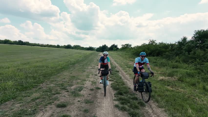A smiling couple rides gravel bicycles along a dirt countryside track during a bikepacking journey through open fields and natural landscapes