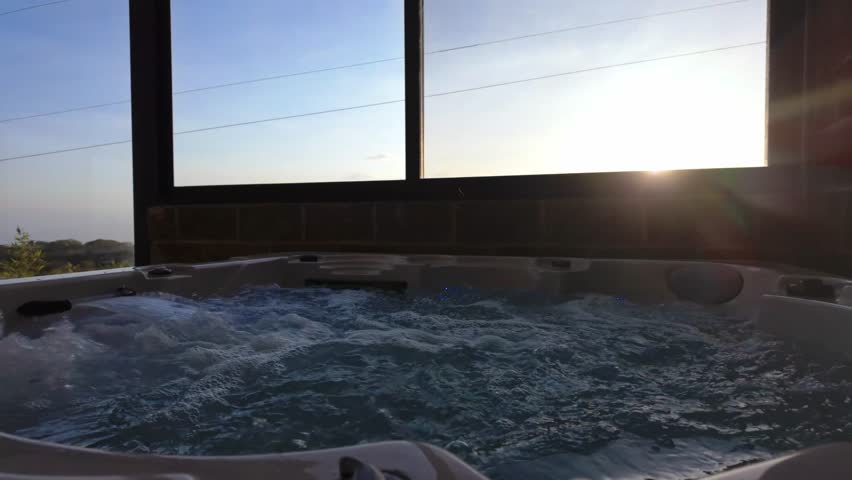 Dolly out shot revealing a bubbling hot tub inside a modern sunroom with large glass windows overlooking a beautiful landscape during a stunning sunset, promoting a concept of relaxation and luxury