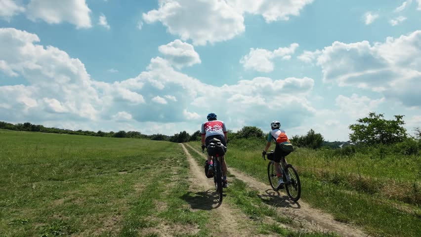 A couple rides gravel bicycles along a dirt track through open countryside landscapes beneath a dramatic sky filled with summer clouds. Rear back view