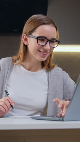 Attractive blonde student in eyeglasses using laptop for studying at home. Woman learn language looks at laptop writes notes on copybook. Slow motion.