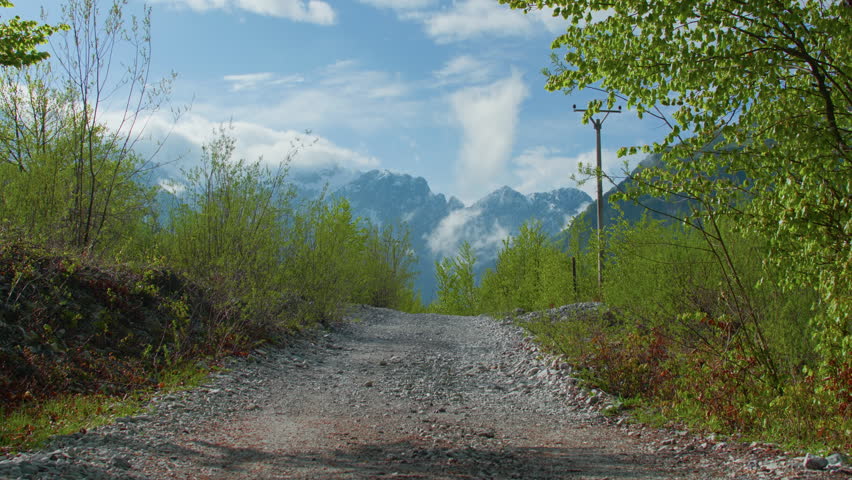 Hiker black backpack gravel path mountains walking away, static tripod shot. Caucasian man in dark gear trekking toward distant peaks through green hills.