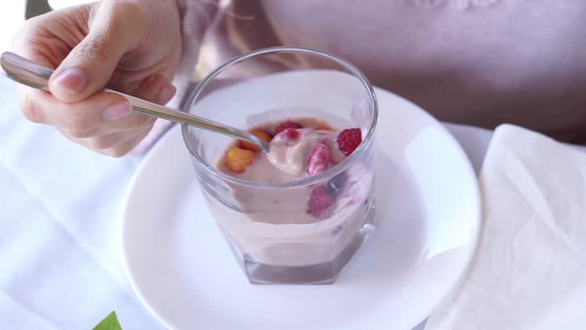 A woman uses a spoon to scoop creamy yogurt mixed with fresh raspberries, blueberries, and cereal from a glass cup during breakfast.
