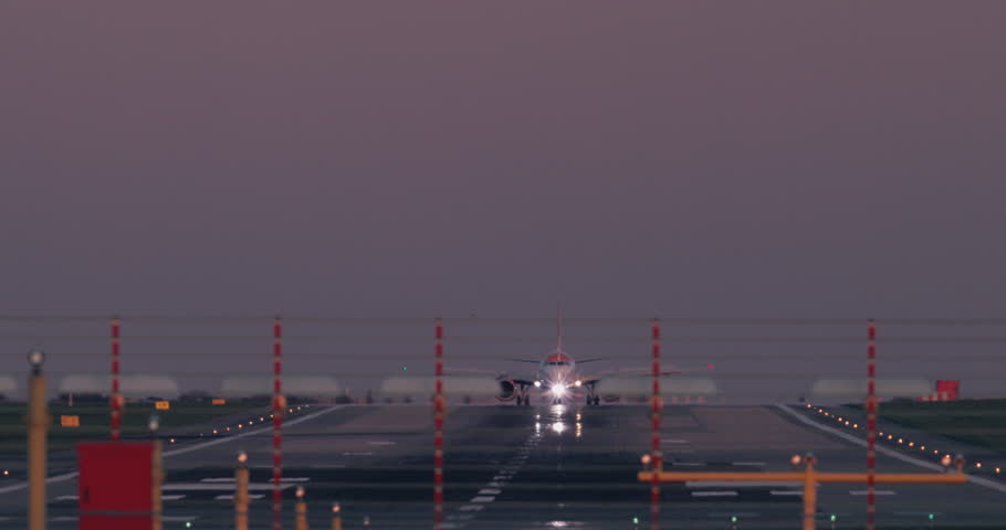 Airplane taxiing on runway at dusk. Front view with bright landing lights, wet surface reflections, airport infrastructure, calm evening atmosphere, aviation ground movement.