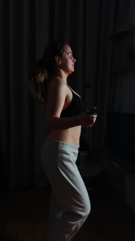 Woman exercising at home in a dimly lit room, focusing on personal fitness and a healthy lifestyle, holding a jump rope handle, working out for weight loss and improved physical well being