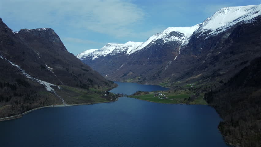 Aerial tilt-down shot descending deeper into a Norwegian fjord valley, revealing a small village with green fields and houses beneath rugged snow-capped mountains.