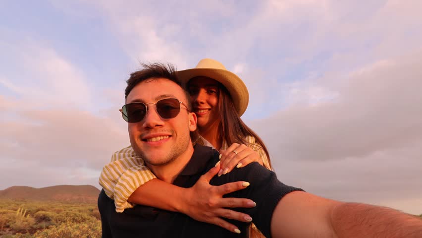 A vibrant couple captures a fun selfie video, laughing and waving while enjoying a playful piggyback ride during a beautiful golden hour in a desert landscape.