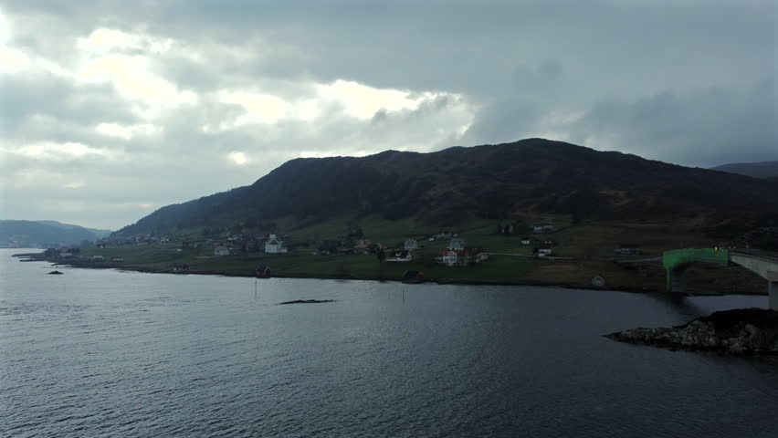Forward push aerial view over a lake toward a hillside with scattered houses and green fields in Norway.