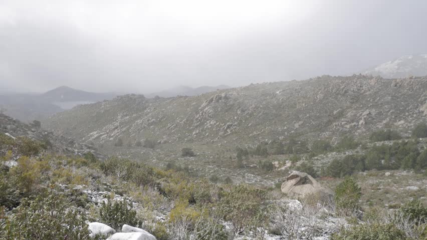 Light snow falls over a rugged mountain landscape with rocky slopes and bushes