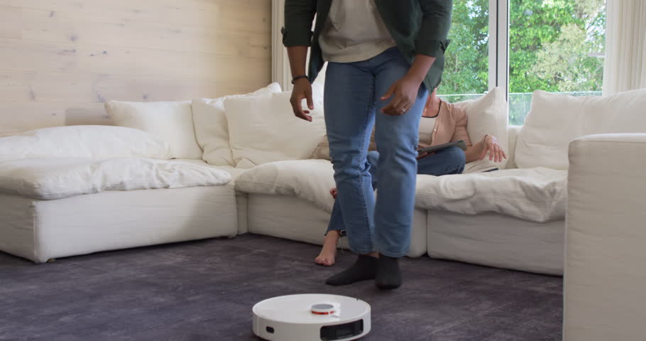 Couple watching robot vacuum moving on dark rug in living room, man crouching, lifting, docking. Cleaner, automated, smart, modern, interior, home, lifestyle